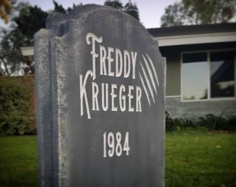 May include: A grey tombstone with the words "Freddy Krueger" and the year "1984" written in white paint. The tombstone is weathered and has a rough texture.