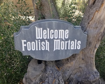 May include: A gray wooden sign with white lettering that reads "Welcome Foolish Mortals". The sign is weathered and has a rustic look.
