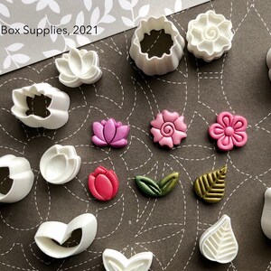 May include: A set of 15 white plastic flower and leaf cookie cutters. The cutters are arranged on a brown patterned surface. The cutters are in the shape of flowers, leaves, and a teardrop. The text "CLAYBOX.CA" is printed on a white plastic sign in the bottom center of the image.