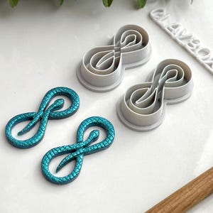 Pode incluir: Dois encantos de resina de cobra infinita azul turquesa e dois cortadores de biscoitos de cobra infinita de plástico branco. Os cortadores de biscoitos estão sobre uma superfície branca. Os encantos estão sobre uma superfície branca.