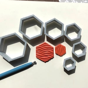 May include: A set of seven grey plastic hexagon-shaped cookie cutters, two orange hexagon-shaped polymer clay pieces with a wavy texture, and a blue and silver tool.