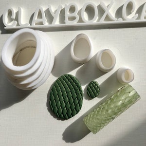Pode incluir: Um conjunto de moldes e ferramentas de argila polimérica para criar joias e outras peças de artesanato. O conjunto inclui uma variedade de formas, incluindo círculos, ovais e um retângulo texturizado. Os moldes são brancos e as ferramentas são verdes.