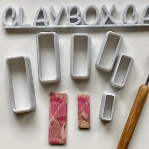Puede incluir: Un juego de seis cortadores de galletas rectangulares de plástico blanco, dos pequeños, dos medianos y dos grandes, con dos baldosas de arcilla polimérica con estampado floral rosa y blanco. Los cortadores de galletas están dispuestos en semicírculo con las baldosas en el centro. El texto "CLAYBOX.CA" es visible en la parte superior de la imagen.