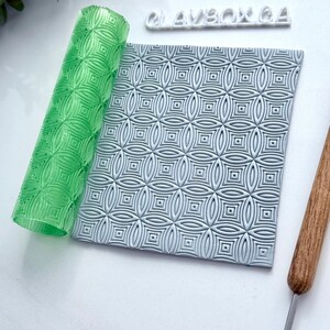 May include: A green patterned rolling pin and a gray patterned clay slab are displayed. The rolling pin is rolled up, showing a repeating geometric design. The clay slab has a similar pattern. The text "CLAYBOX 64" is above the clay slab.