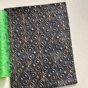 May include: A black and gold textured rolling pin with a repeating pattern of swirls and diamonds. The rolling pin is made of a flexible material and is designed for use with fondant or other edible materials.