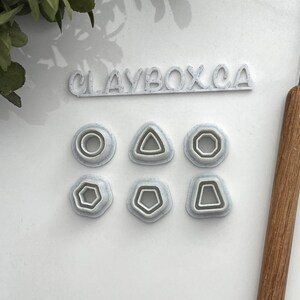 May include: Set of six white plastic cookie cutters with different shapes: circle, triangle, hexagon, star, pentagon, and a rounded rectangle. The cutters are on a white surface with the text "CLAYBOX.CA" above them.