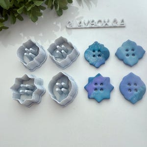 May include: Four plastic cookie cutters with a star, flower, and two button shapes.  Next to the cutters are four clay buttons in the same shapes, with blue, purple, and white marbled designs.