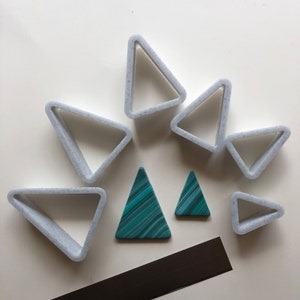 May include: An overhead shot shows a collection of geometric shapes. Several light grey triangle-shaped cookie cutters are scattered around two teal and green triangle-shaped objects. A dark grey rectangular magnet is also present.