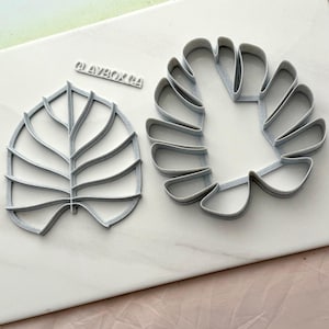 May include: Two gray plastic leaf-shaped cookie cutters. One cutter is a detailed leaf with veins, and the other is a stylized leaf with rounded edges. The text "@CLAYBOX.CA" is visible in the background.