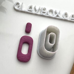 May include: Two pink plastic cookie cutters shaped like the letter "o" with a textured surface. The larger cutter is nested inside the smaller cutter.