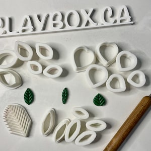 Puede incluir: Un conjunto de cortadores de galletas de plástico blanco en varias formas, incluyendo hojas, corazones y óvalos. También hay tres piezas de arcilla polimérica en forma de hoja verde y una herramienta de madera con un gancho de metal.