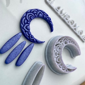 Op de afbeelding: Een close-up van een verzameling knutselgereedschap. Een donkerblauw, halvemaanvormig object met een gedetailleerd patroon ligt naast een lichtgrijze, halvemaanvormige uitsteker. Er zijn ook drie blauwe ovale vormen en een wit object met de tekst "AVEOX".