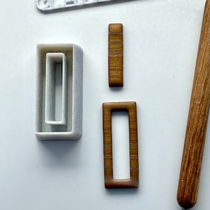 Puede incluir: Tres cortadores de galletas de madera en forma de rectángulos con esquinas redondeadas. Los cortadores son de color marrón con un patrón de veta de madera. El cortador más grande mide aproximadamente 5 cm de largo y 2 cm de ancho. El cortador del medio mide aproximadamente 4 cm de largo y 1,5 cm de ancho. El cortador más pequeño mide aproximadamente 3 cm de largo y 1 cm de ancho.