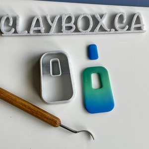 May include: White plastic clay cutters in the shape of a rectangle and a mouse. A blue and green gradient clay rectangle with a hole in the centre. A blue clay rectangle. A wooden tool with a metal hook.