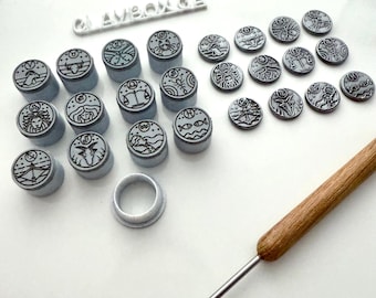 Op de afbeelding: Een verzameling grijze en zwarte dierenriemstempels en ronde afdrukken. De stempels zijn cilindrisch met een dierenriemsymbool erop. Een gereedschap met houten handvat en een ring zijn ook aanwezig.