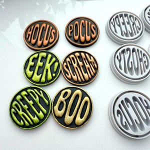 May include: A collection of Halloween-themed round decorations. The decorations feature words like "Hocus," "Pocus," "Eek," "Scream," "Creepy," and "Boo" in a stylized font. The color palette includes orange, green, and gold.
