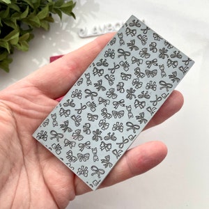 Pode incluir: Uma placa de embossing de metal com um padrão repetitivo de pequenos laços. A placa é de cor prateada e tem uma forma retangular.