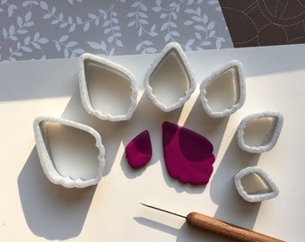 May include: A set of white plastic cookie cutters in various flower shapes, including a butterfly, a heart, and a leaf. There are also two pieces of purple polymer clay, one shaped like a butterfly and the other a heart. A wooden tool is also included.