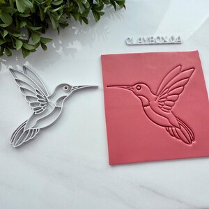 May include: A white hummingbird-shaped cookie cutter and a pink rectangular stamp with a hummingbird design. The stamp has the text "CLAYBOXOA" on it. The items are on a white surface.