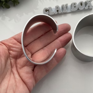 Könnte beinhalten: Zwei silberne Metall-Armreifen, einer ist offen und wird in einer Hand gehalten, der andere ist geschlossen und liegt auf einer weißen Oberfläche. Das Wort "CLAY BOX" ist im Hintergrund sichtbar.