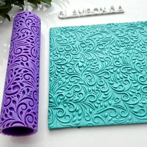May include: A purple cylindrical rolling pin and a flat rectangular sheet, both featuring an intricate embossed swirling vine and leaf pattern. The text CLAYBOX.CA is visible in white lettering near the top of the composition.