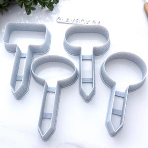 May include: Four light blue, T-shaped cookie cutters in varying sizes. Each cutter has a rounded top and a handle with a pointed end. The cutters are made of plastic and are arranged on a white surface.