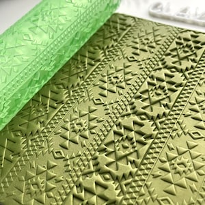 Puede incluir: Rodillo verde con un patrón geométrico, utilizado para crear diseños en relieve en fondant o arcilla. El rodillo se muestra sobre una superficie verde con el mismo patrón.