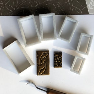May include: A set of seven white plastic rectangular frames, some with rounded corners, and two brass rectangular stamps with intricate designs. The frames are arranged on a white surface.