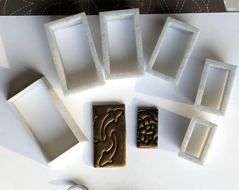 May include: A set of seven white plastic rectangular frames, some with rounded corners, and two brass rectangular stamps with intricate designs. The frames are arranged on a white surface.