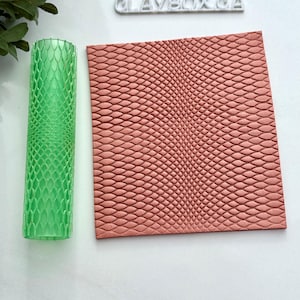 Peut inclure: Un rouleau texturé cylindrique vert et une feuille de texture d'argile rectangulaire couleur saumon. Le rouleau présente un motif en forme d'écailles et la feuille présente un motif en relief similaire. Le texte "CLAYBOX CA" est visible en arrière-plan.