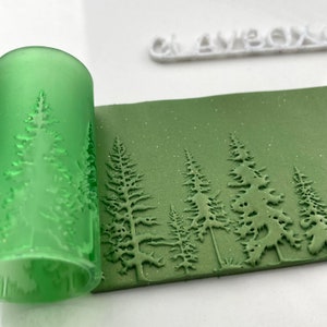 Peut inclure: Un rouleau à pâtisserie vert avec un motif en relief d'arbres de pin est utilisé pour créer une empreinte dans une surface en pâte polymère verte. La pâte a une finition scintillante.