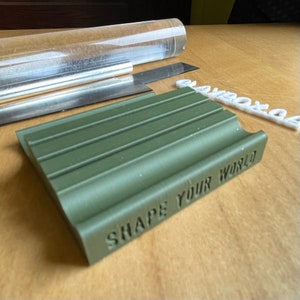 May include: An olive green rectangular object with the text "SHAPE YOUR WORLD" is in the foreground. A clear acrylic roller, metal tools, and a white plastic object with the word "somboxe" are also visible on a wooden surface.