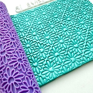 May include: A purple cylindrical rolling pin and a rectangular teal flat sheet, both featuring a repeating embossed daisy flower pattern. The rolling pin has a raised floral design, while the flat sheet shows the same daisy pattern pressed into it.