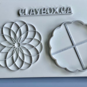 Peut inclure: Deux emportes-pièces en plastique blanc, l'un avec un motif circulaire et l'autre avec un motif en forme de fleur. Le texte "CLAYBOX.CA" est imprimé sur la surface.