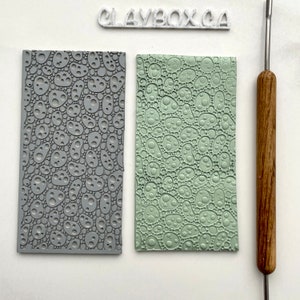 May include: Two polymer clay texture sheets, one grey and one green, with a repeating pattern of small circles. The sheets are on a white surface next to a wooden sculpting tool.
