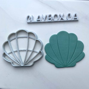 Puede incluir: Un cortador de galletas de plástico blanco en forma de concha y una concha de arcilla verde. El cortador de galletas tiene un diseño detallado con crestas. La concha de arcilla es lisa y tiene un diseño ligeramente elevado.