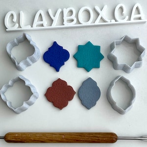 May include: A set of eight white plastic cookie cutters in various shapes, including a star, a circle, and a quatrefoil. The cutters are arranged on a white surface with a wooden rolling pin and a sign that reads "CLAYBOX.CA".
