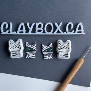 May include: Four white plastic cookie cutters shaped like mummified cats with green eyes. The cutters are on a black surface with the text "CLAYBOX.CA" above them.