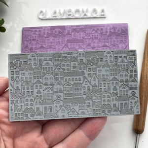 May include: A gray rubber stamp with a detailed design of a city skyline. The stamp is rectangular and has a raised design of buildings with windows and roofs. The stamp is labeled "Claybox CA" in white lettering.