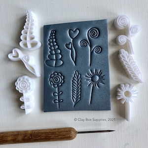 Puede incluir: Un conjunto de 9 sellos de arcilla polimérica blanca con diseños florales. Los sellos están dispuestos alrededor de una lámina de arcilla polimérica gris con los mismos diseños florales tallados en ella. La lámina tiene el texto "Clay Box Supplies, 2021" impreso en ella. También se incluye una herramienta de madera con una punta de metal.