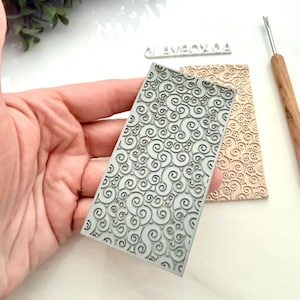 Puede incluir: Un sello rectangular de textura de arcilla con un patrón de remolino se sostiene en una mano. Un segundo sello con el mismo diseño está al lado. También se ve una herramienta de madera. Los sellos son para arte y artesanía en arcilla.