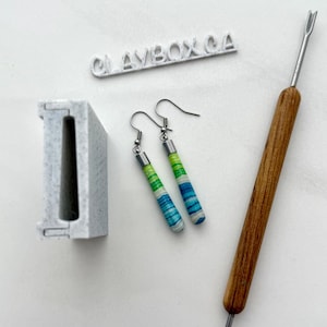 May include: Cylindrical earrings in green, blue, and white, with silver hooks. The image also shows a clay tool and a grey rectangular object. The text "CLAYBOX CA" is visible. The earrings are a handmade craft item.