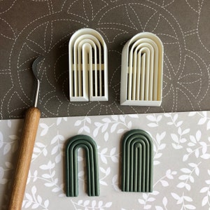 Puede incluir: Dos moldes de arco de plástico blanco y dos arcos de arcilla polimérica verde. Los moldes tienen varias capas para crear una forma de arco tridimensional. Los arcos de arcilla son un producto terminado.
