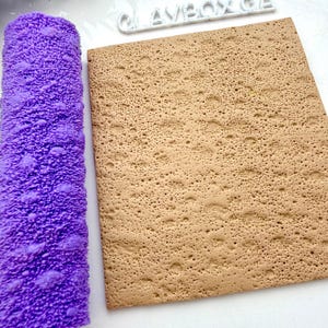Puede incluir: Un rodillo morado texturizado y una lámina rectangular marrón con un patrón punteado. El rodillo es cilíndrico con una superficie irregular. La lámina tiene una textura rugosa y porosa. La palabra "CLAYBOX" es visible en blanco.