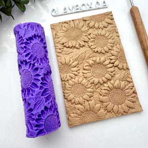 May include: A purple rolling pin with a sunflower pattern and a tan clay slab with a sunflower design. The rolling pin is cylindrical and has a repeating sunflower pattern. The clay slab has a raised sunflower design. A tool with a wooden handle is also visible.