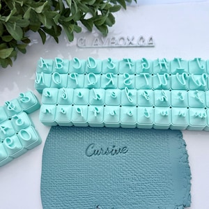 May include: A collection of light blue polymer clay letter and number stamps, with a matching blue clay sheet that reads "Cursive". The stamps are arranged in rows, with the letters and numbers clearly visible. The brand name "CLAYBOX.CA" is visible in the background.