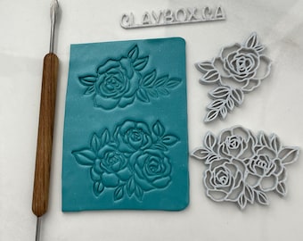 Puede incluir: Un conjunto de tres moldes de arcilla polimérica blanca con un diseño floral de rosas y hojas. Los moldes están sobre una estera de silicona azul turquesa. También se incluye una herramienta de madera con una punta de metal.