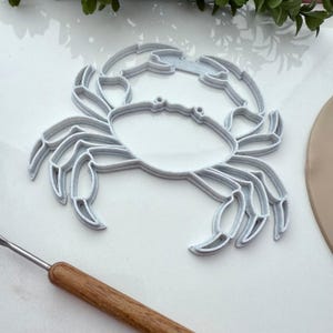 May include: A white 3D printed crab cookie cutter. The cutter is made of plastic and has a detailed design.