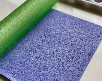 May include: A close-up of a light purple textured fondant sheet with a green rolling pin on top of it.