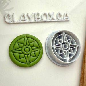 Può includere: Un disegno di rosa dei venti verde in argilla, accanto a un tagliabiscotti in plastica grigia con lo stesso disegno. La parola "CLAYBOX CA" è sopra gli oggetti. La rosa dei venti è un disegno circolare.